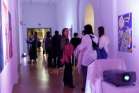 Besucherinnen und Besucher betrachten bunte Kunstwerke an den Wänden eines Ausstellungsflurs mit lila Beleuchtung.