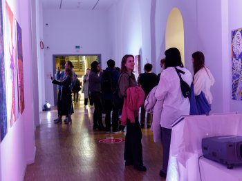 Foto: Lukas Pürmayr
Besucherinnen und Besucher betrachten bunte Kunstwerke an den Wänden eines Ausstellungsflurs mit lila Beleuchtung.