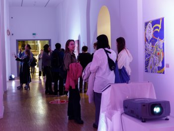 Besucherinnen und Besucher betrachten bunte Kunstwerke an den Wänden eines Ausstellungsflurs mit lila Beleuchtung.