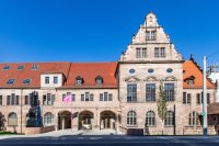 Das Künstlerhaus mit rotem Ziegeldach, verziertem Giebel und Bogeneingang unter blauem Himmel, davor weht eine pinke Fahne.