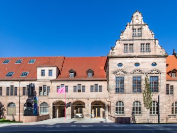 Foto: Dieter Zeitler
Das Künstlerhaus mit rotem Ziegeldach, verziertem Giebel und Bogeneingang unter blauem Himmel, davor weht eine pinke Fahne.