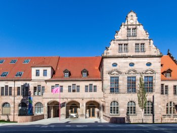 Das Künstlerhaus mit rotem Ziegeldach, verziertem Giebel und Bogeneingang unter blauem Himmel, davor weht eine pinke Fahne.
