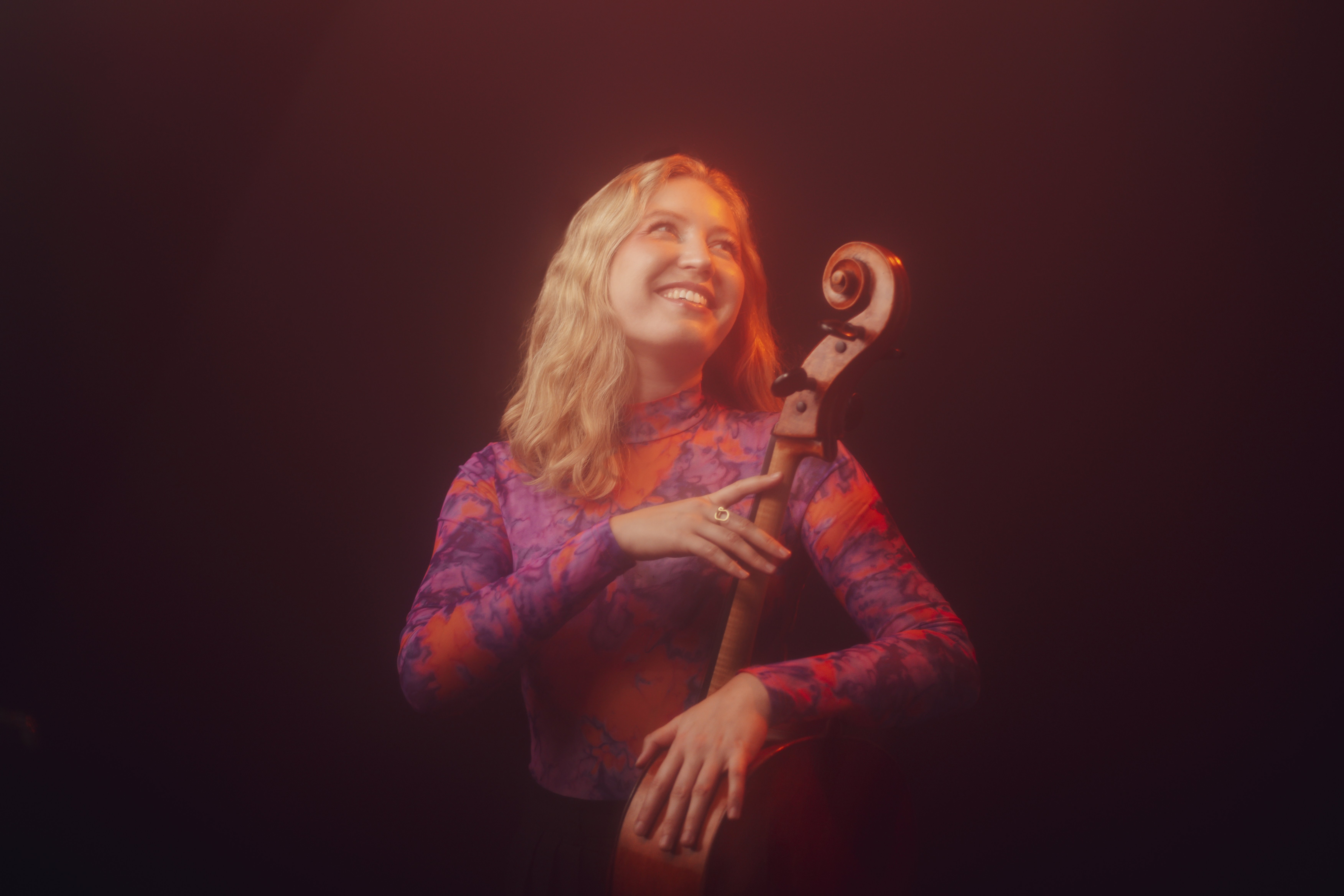 Lachende Person mit blonden Haaren lehnt sich an ein Violoncello, in warmes rötliches Licht getaucht.