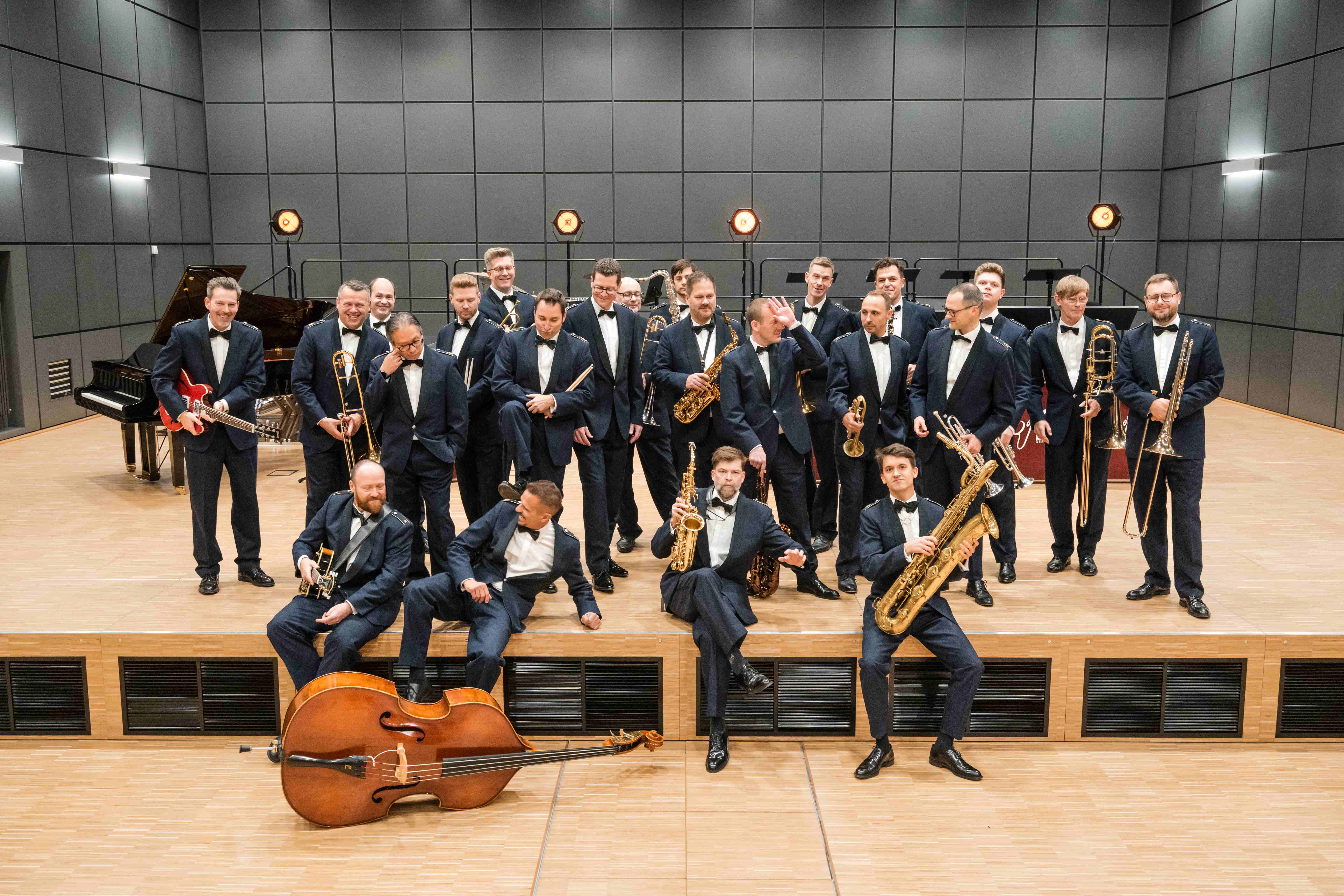 Eine Big Band in dunklen Anzügen posiert mit Blasinstrumenten, Gitarren und einem Kontrabass auf einer Bühne mit Klavier im Hintergrund.