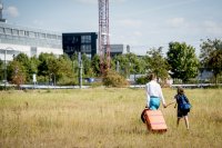 Melli (Franziska Hartmann) und ihr 9-jähriger Sohn Ben (Claudio Magno) gehen händehaltend mit einem orangefarbenen Rollkoffer über eine Wiese auf Gebäude im Hintergrund zu.