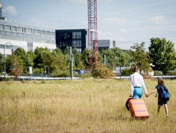 Copyright: © 2Pilots Filmproduction
Melli (Franziska Hartmann) und ihr 9-jähriger Sohn Ben (Claudio Magno) gehen händehaltend mit einem orangefarbenen Rollkoffer über eine Wiese auf Gebäude im Hintergrund zu.