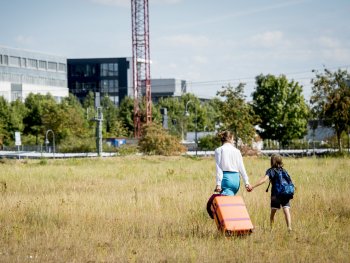 Copyright: © 2Pilots Filmproduction Melli (Franziska Hartmann) und ihr 9-jähriger Sohn Ben (Claudio Magno) gehen händehaltend mit einem orangefarbenen Rollkoffer über eine Wiese auf Gebäude im Hintergrund zu.