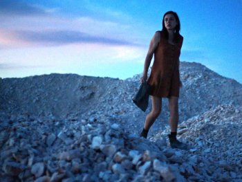 Copyright: NSFA
Eine Frau in kurzem Kleid steht mit einer Tasche in der Hand auf einem Schutthügel aus hellem Gestein in der Abenddämmerung.
