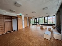 Leerer Raum mit Parkettboden, großen Fenstern, Wandschränken und mehreren kleinen Holzhockern mit gepolsterten Sitzflächen.