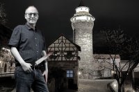 Ein Mann mit Brille und Messer in der Hand steht bei Nacht vor einem beleuchteten Turm und einem Fachwerkhaus.