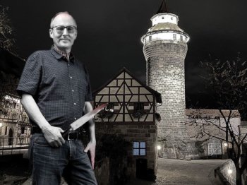 Ein Mann mit Brille und Messer in der Hand steht bei Nacht vor einem beleuchteten Turm und einem Fachwerkhaus.