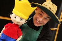 Ein lächelnder Puppenspieler mit Strohhut hält eine Handpuppe mit gelber Krone, rotem Oberteil und blauer Hose neben seinem Gesicht.