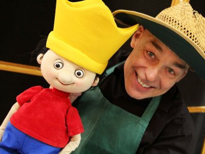 Ein lächelnder Puppenspieler mit Strohhut hält eine Handpuppe mit gelber Krone, rotem Oberteil und blauer Hose neben seinem Gesicht.