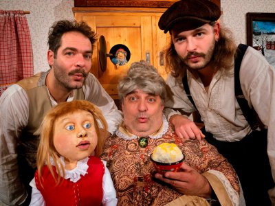 Drei Schauspieler in historischen Kostümen posieren mit einer Handpuppe und einer Schale Speiseeis vor einem Holzschrank mit kleiner Wichtelfigur.