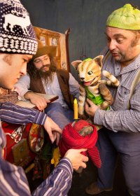 Schauspieler in Kostümen präsentieren bei einer Theaterszene eine Katzen-Handpuppe und einen in rote Wolle gewickelten Gegenstand mit Kerze.