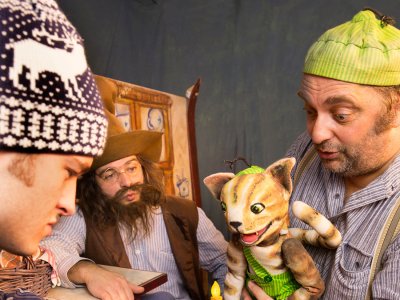 Schauspieler in Kostümen präsentieren bei einer Theaterszene eine Katzen-Handpuppe und einen in rote Wolle gewickelten Gegenstand mit Kerze.