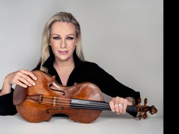 Eine Frau mit blonden Haaren in schwarzer Kleidung hält lächelnd eine Geige vor sich auf einem Tisch.