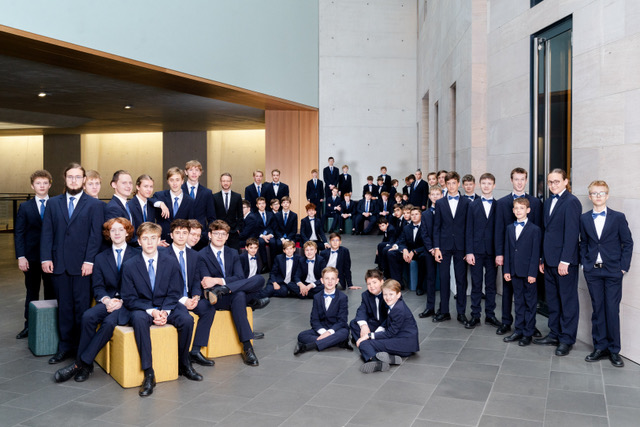 Eine große Gruppe von Jungen und jungen Männern in dunklen Anzügen mit Fliegen posiert für ein Gruppenfoto in einer modernen Halle.