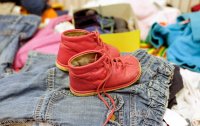 Ein Paar rote Kinderschuhe steht auf einem Haufen gebrauchter Kleidungsstücke, im Hintergrund weitere Second-Hand-Kleidung unscharf zu sehen.