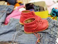 Ein Paar rote Kinderschuhe steht auf einem Haufen gebrauchter Kleidungsstücke, im Hintergrund weitere Second-Hand-Kleidung unscharf zu sehen.