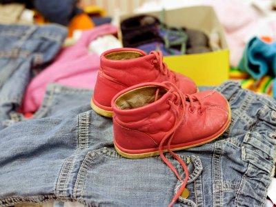 Ein Paar rote Kinderschuhe steht auf einem Haufen gebrauchter Kleidungsstücke, im Hintergrund weitere Second-Hand-Kleidung unscharf zu sehen.