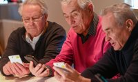 Drei ältere Männer sitzen nebeneinander an einem Tisch und betrachten gemeinsam mehrere Smartphones in ihren Händen.