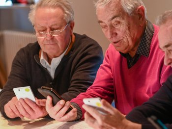 Drei ältere Männer sitzen nebeneinander an einem Tisch und betrachten gemeinsam mehrere Smartphones in ihren Händen.