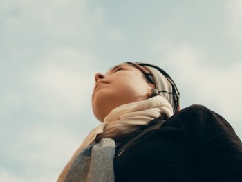 Person mit Kopftuch blickt vor blauem Himmel mit Wolken nach oben.