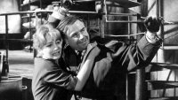Schwarz-Weiß-Filmszene, in der ein Mann mit gefesselten Handgelenken eine ängstlich wirkende Frau in einer Industrieanlage schützend festhält.