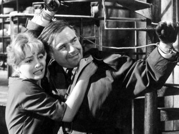 Copyright: Filmverleih Schwarz-Weiß-Filmszene, in der ein Mann mit gefesselten Handgelenken eine ängstlich wirkende Frau in einer Industrieanlage schützend festhält.