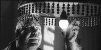 Schwarz-weißes Filmstill zeigt eine Person mit langen Fingernägeln, die unter einem mit Perlenketten verzierten Lampenschirm nach einer leuchtenden Glühbirne greift.