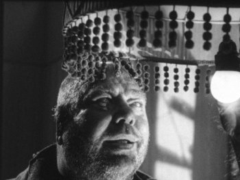 Copyright: Filmverleih
Schwarz-weißes Filmstill zeigt eine Person mit langen Fingernägeln, die unter einem mit Perlenketten verzierten Lampenschirm nach einer leuchtenden Glühbirne greift.