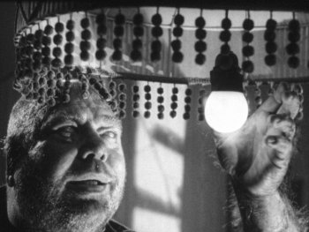 Copyright: Filmverleih Schwarz-weißes Filmstill zeigt eine Person mit langen Fingernägeln, die unter einem mit Perlenketten verzierten Lampenschirm nach einer leuchtenden Glühbirne greift.