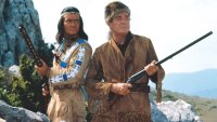 Zwei Männer in traditioneller Lederkleidung stehen mit Gewehren in einer felsigen Landschaft.