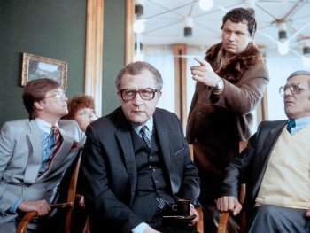 Copyright: Filmverleih
Mehrere Personen in Anzügen sitzen und stehen in einem Raum, während eine stehende Person mit Pelzkragen-Mantel gestikuliert.