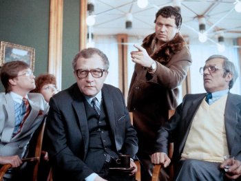 Copyright: Filmverleih Mehrere Personen in Anzügen sitzen und stehen in einem Raum, während eine stehende Person mit Pelzkragen-Mantel gestikuliert.