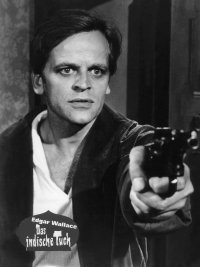 Schwarz-Weiß-Filmszene aus „Edgar Wallace – Das indische Tuch“, in der ein Mann bedrohlich eine Pistole auf die Kamera richtet.