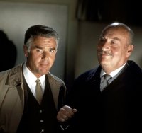 Zwei Männer im Anzug stehen nah beieinander in einem abgedunkelten Raum, einer trägt einen hellen Trenchcoat, der andere einen dunklen Samtmantel.