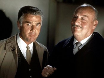 Copyright: Filmverleih Zwei Männer im Anzug stehen nah beieinander in einem abgedunkelten Raum, einer trägt einen hellen Trenchcoat, der andere einen dunklen Samtmantel.