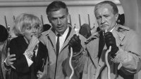 Schwarz-weiß Filmszene mit einer besorgten Frau und zwei Männern in Trenchcoats, die Kabelbinder oder Schnürsenkel in den Händen halten.