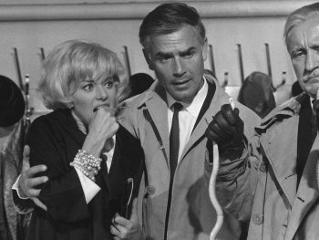 Copyright: Filmverleih
Schwarz-weiß Filmszene mit einer besorgten Frau und zwei Männern in Trenchcoats, die Kabelbinder oder Schnürsenkel in den Händen halten.