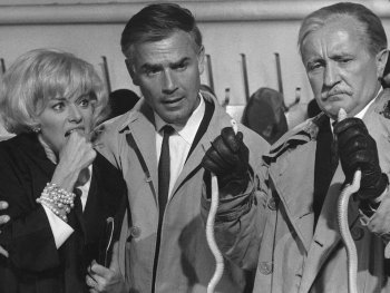 Copyright: Filmverleih Schwarz-weiß Filmszene mit einer besorgten Frau und zwei Männern in Trenchcoats, die Kabelbinder oder Schnürsenkel in den Händen halten.
