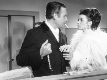 Copyright: Filmverleih Schwarz-Weiß-Filmszene: Ein Mann im Anzug mit Zigarette hält eine Halskette und steht einer Frau in einem Federmantel gegenüber, die ihn ansieht.