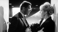 Schwarzweißfoto einer Filmszene mit einem Mann im Anzug und einer Frau mit hochgestecktem Haar, die sich in angespanntem Gespräch gegenüberstehen.