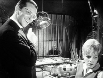 Copyright: Filmverleih Schwarz-weiß Filmszene mit einem grinsenden Mann mit Brille und einer besorgt blickenden Frau in einem Labor mit Operationsliege und einem Affen im Käfig im Hintergrund.