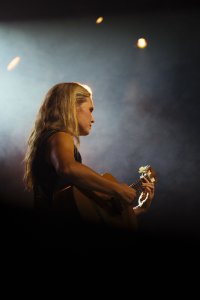 Person mit langen blonden Haaren spielt im Profil auf einer Bühne konzentriert Gitarre, im Hintergrund verschwommene Bühnenlichter.
