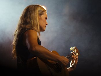 Person mit langen blonden Haaren spielt im Profil auf einer Bühne konzentriert Gitarre, im Hintergrund verschwommene Bühnenlichter.