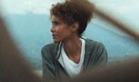 Person mit lockigem Haar blickt zur Seite, im Hintergrund eine hügelige Landschaft im Dunst.