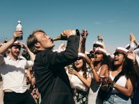 Ein Mann trinkt aus einer Flasche, während ihn eine jubelnde Gruppe in weißen Studentenmützen umgibt.