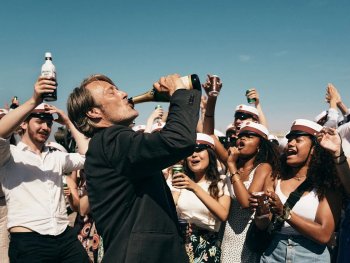 Copyright: Weltkino Filmverleih
Ein Mann trinkt aus einer Flasche, während ihn eine jubelnde Gruppe in weißen Studentenmützen umgibt.